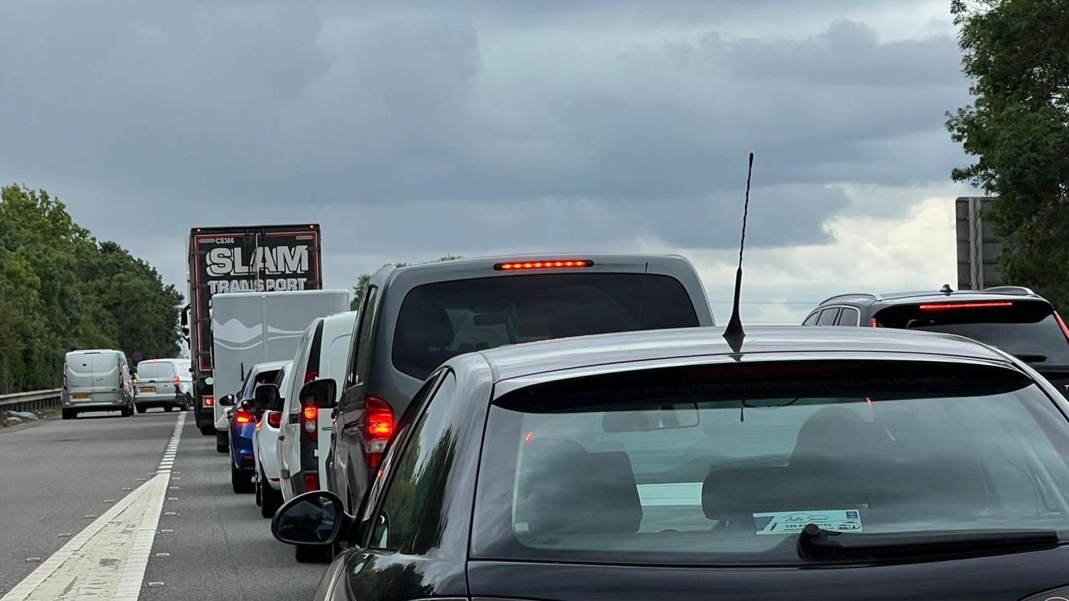 The dual carriageway of the A1 where a slip road is merging on to the carriageway. There are cars and lorries queuing.