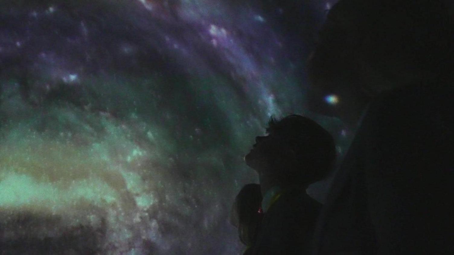 Three students in the mobile planetarium looking up at a green and purple solar system