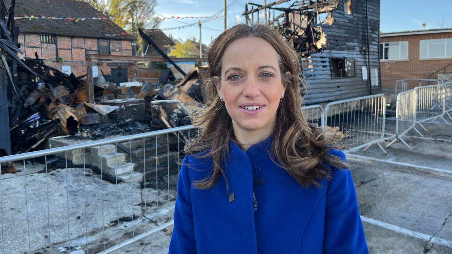 A blonde woman in a dark blue coat. She is standing in front of a rubble of burnt buildings squared off by metal fencing.