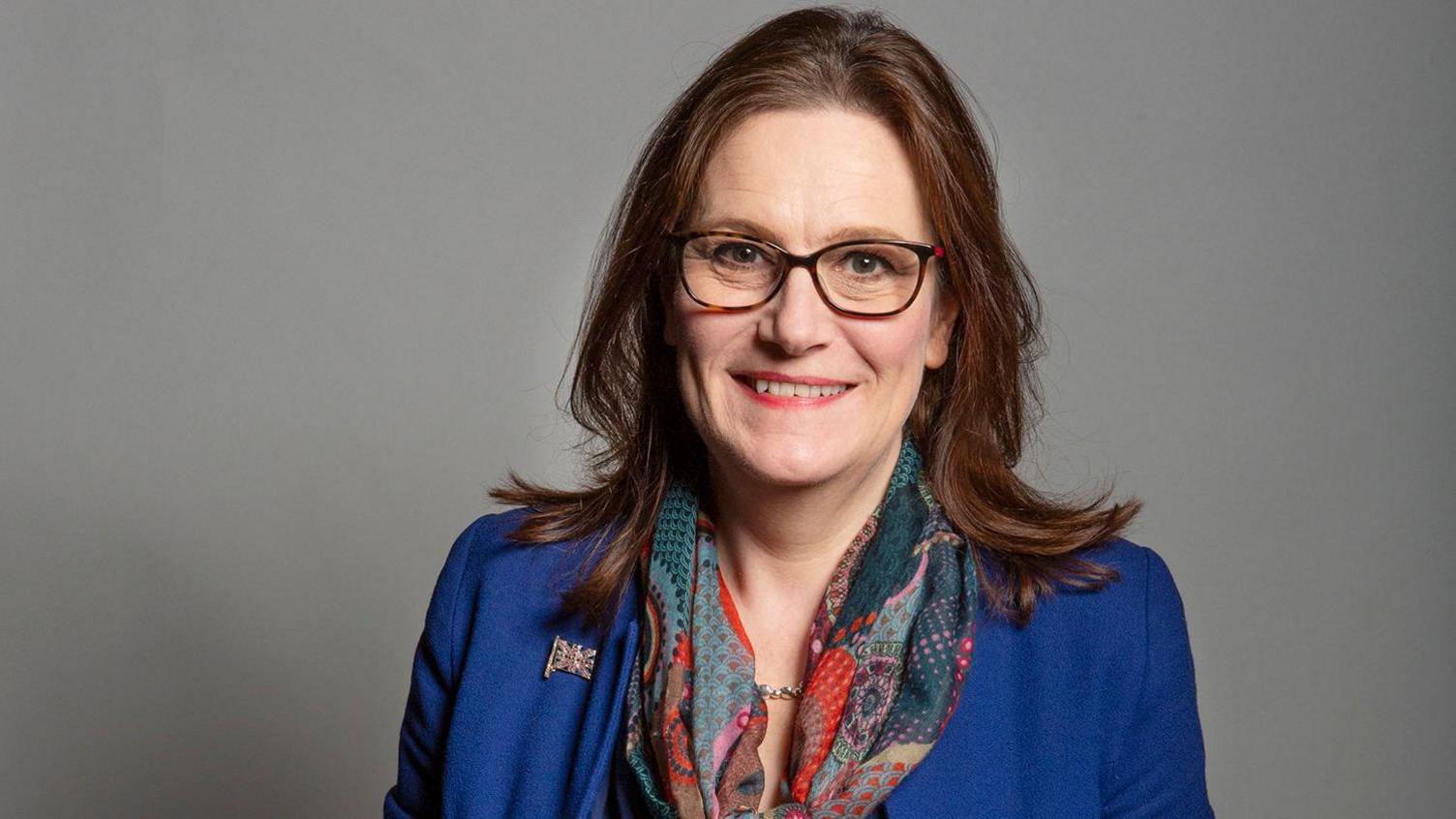 Rebecca Harris smiles into the camera. She is wearing a blue blazer, colourful scarf, and glasses.