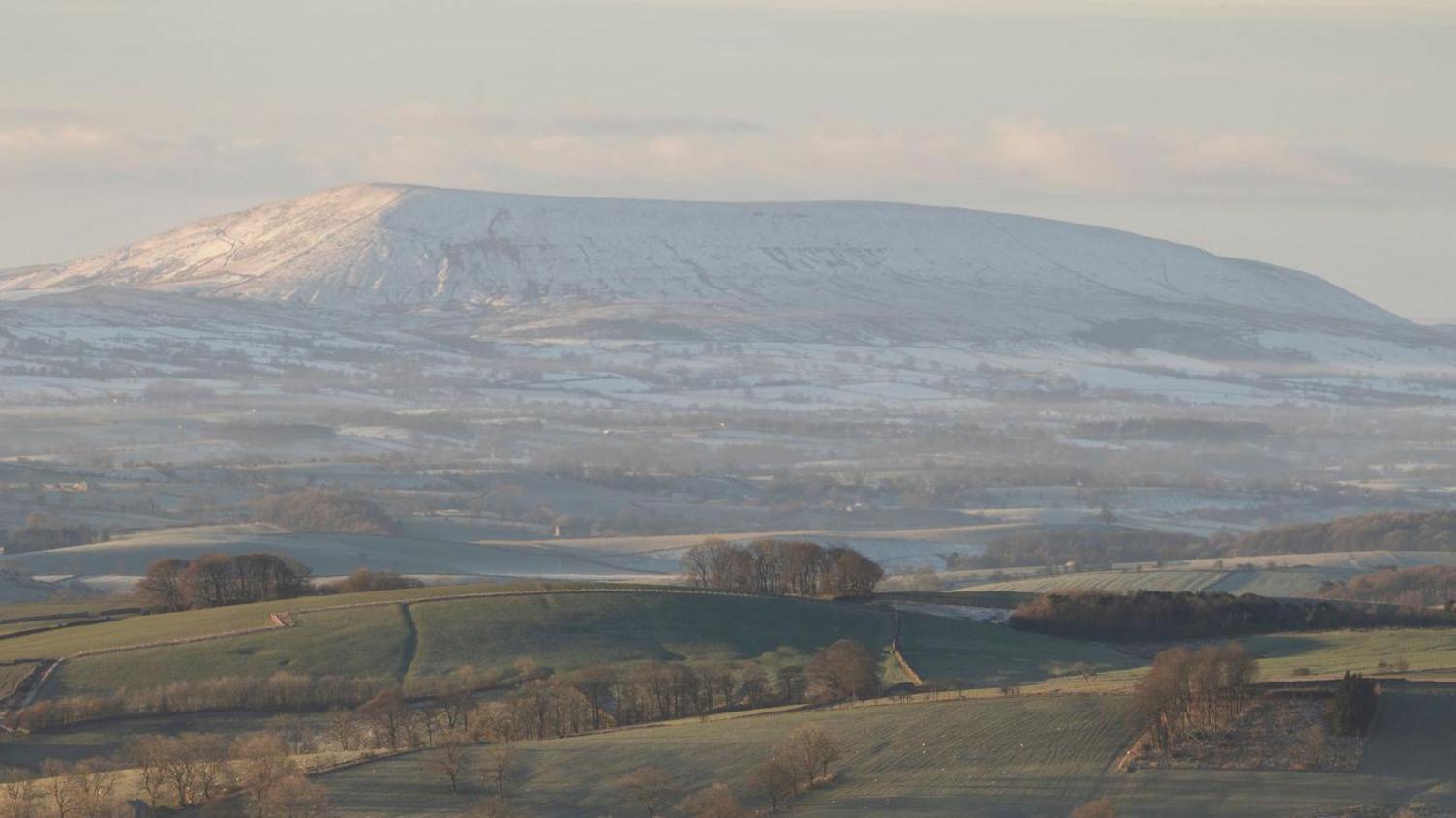 Snow shrouds Pendle Hill with green slopes and fields in the foreground