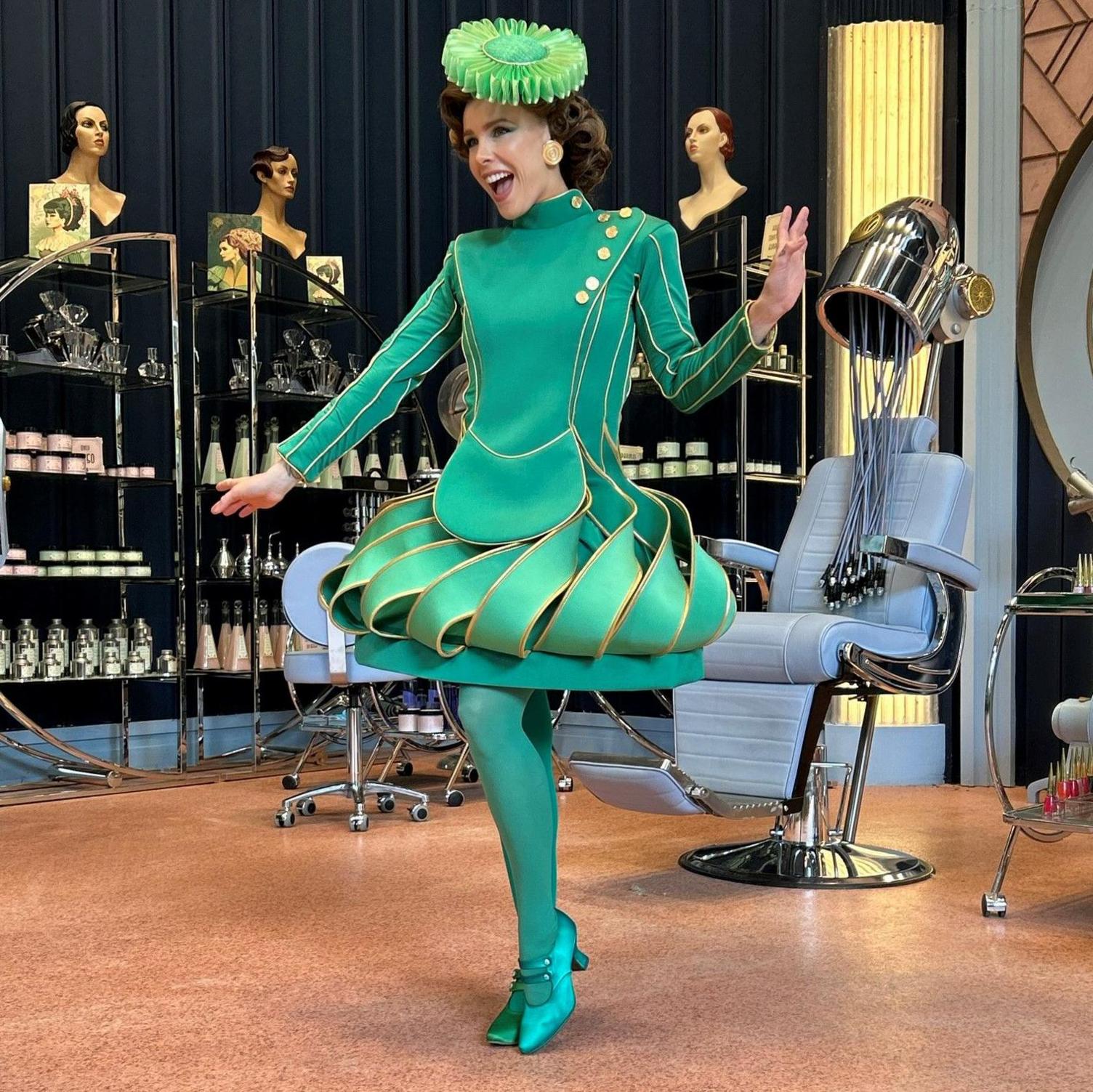 Lily Field wearing an Emerald City costume in the film Wicked. She is striking a pose in a scene in a hairdressing salon.