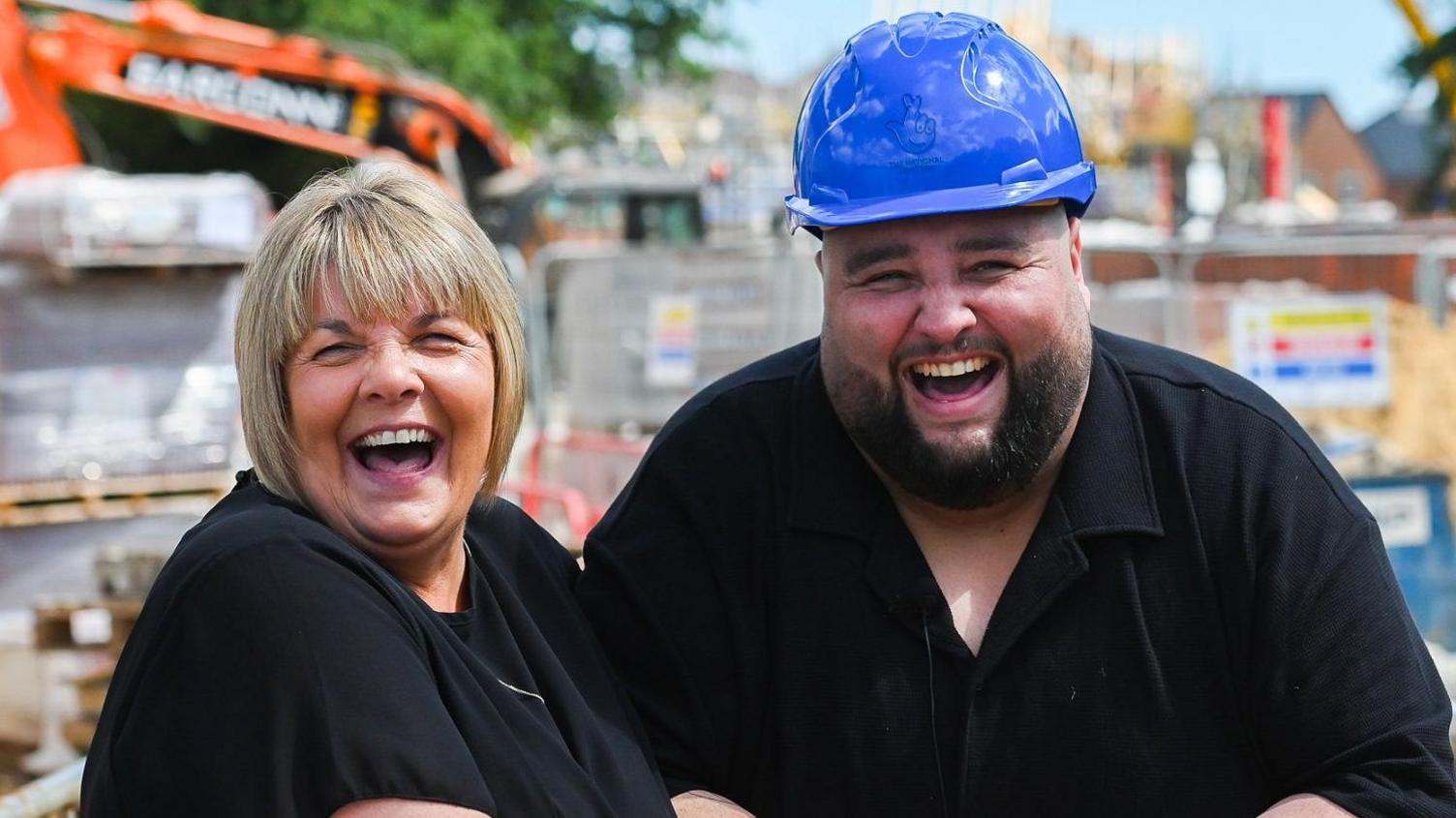 Dannica and Adam Lopez standing outside at a building site. Dannica is a woman with a blonde bob who is looking at the camera and laughing, she is wearing a black T-shirt. Next to her, on the right, Adam Lopez is wearing a blue hard hat and is wearing a black T-shirt. He is looking directly at the camera and laughing.