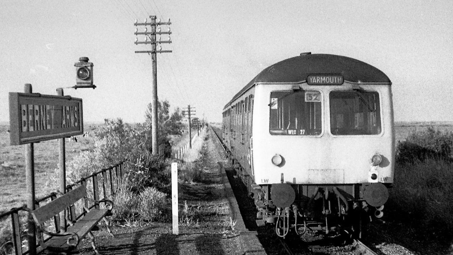 A black and white image of an old train on a railway line running through a field. A sign on the front of the train reads 'Yarmouth'. To the left of the railway line there is a bench with a sign above it that reads 'Berney Arms'.