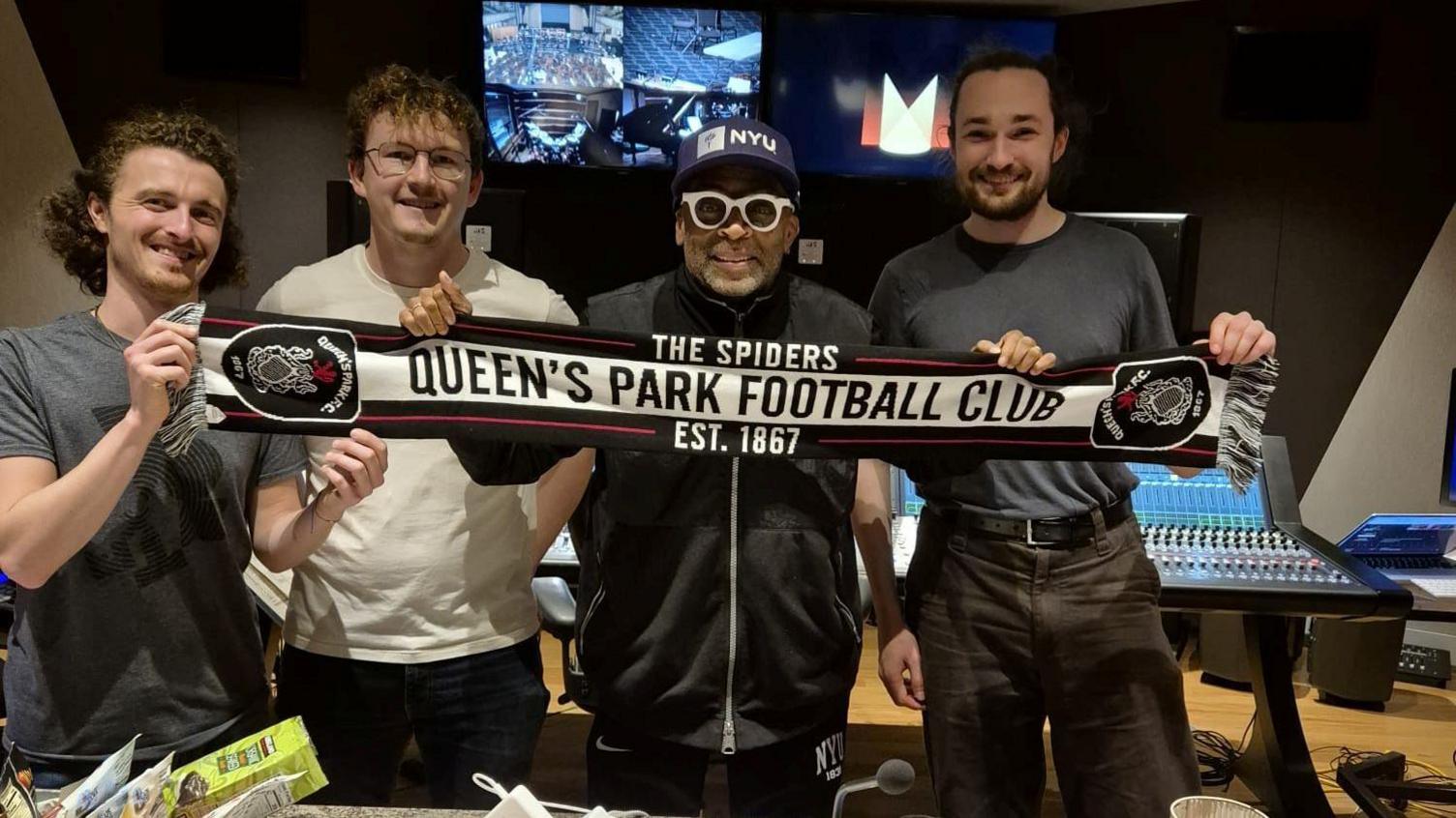 Four men standing in a recording studio. They are holding a scarf saying Queen's Park Football Club on it.