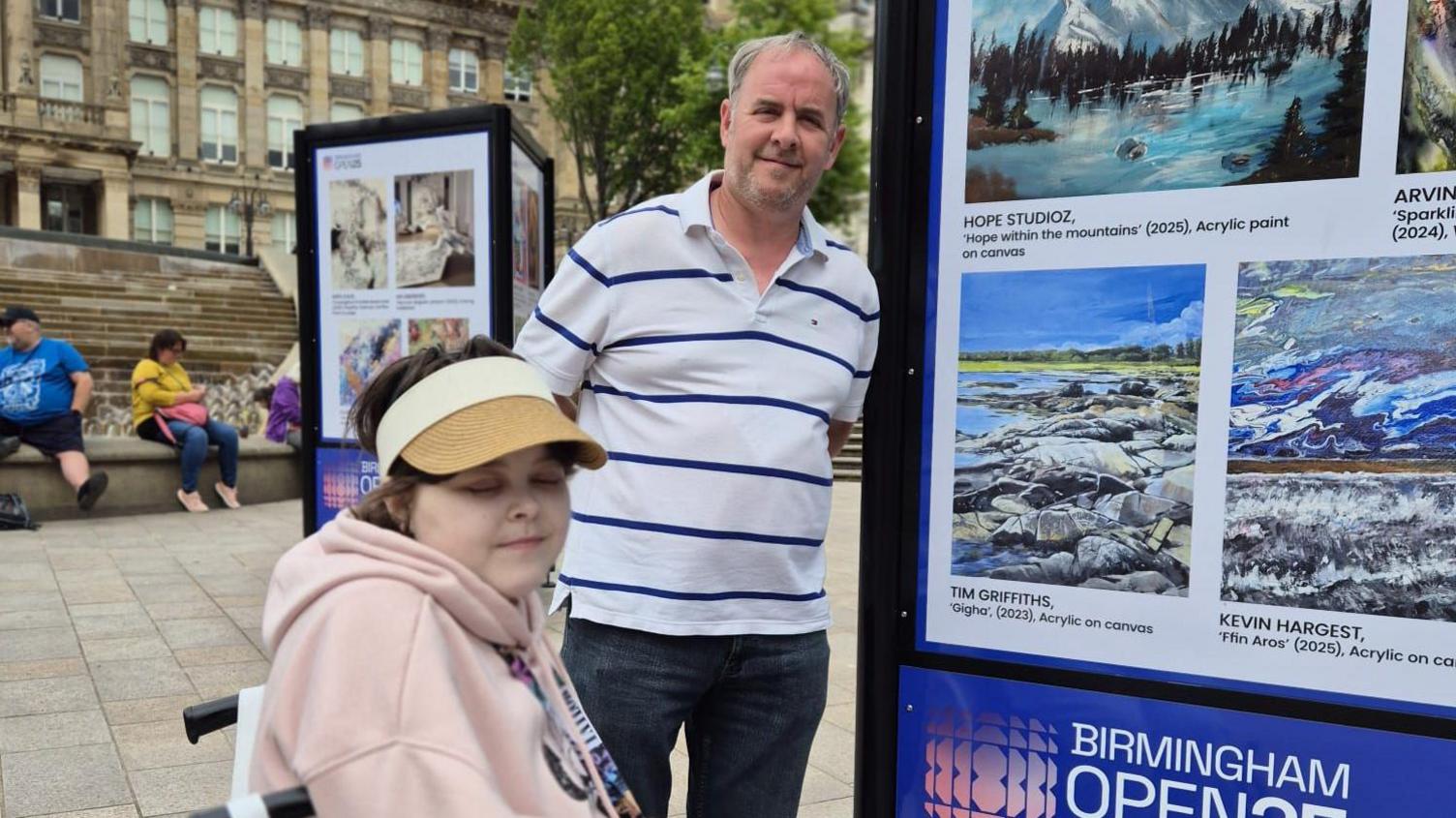 Lily with her dad Graham. They are standing next to her artwork. She has a beige Micky Mouse hoody on and a beige hat. He has a white and blue polo shirt on.