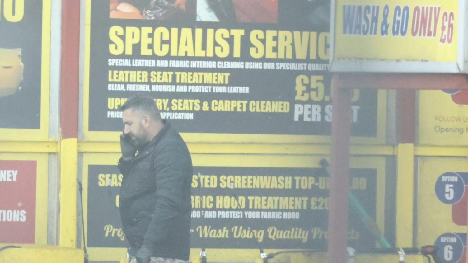 Khdir in a black coat and is on the phone. He is in a car wash where there are yellow signs offering differnt car wash services.