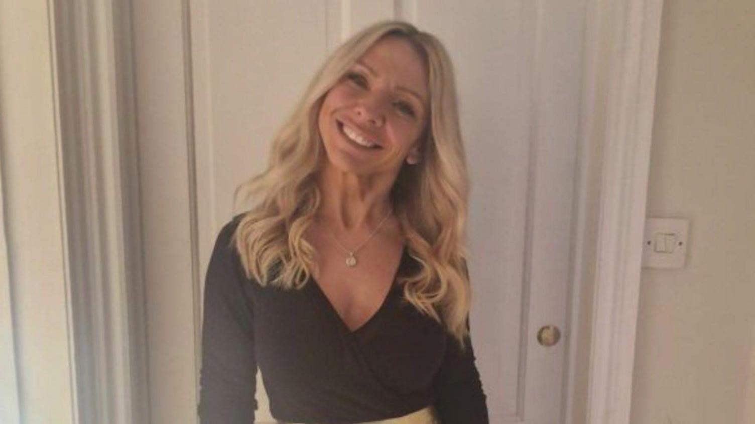 Claire Chick seen with long wavy blonde hair and smiling at the camera. She is wearing a black top with a gold skirt in front of a white doorway.