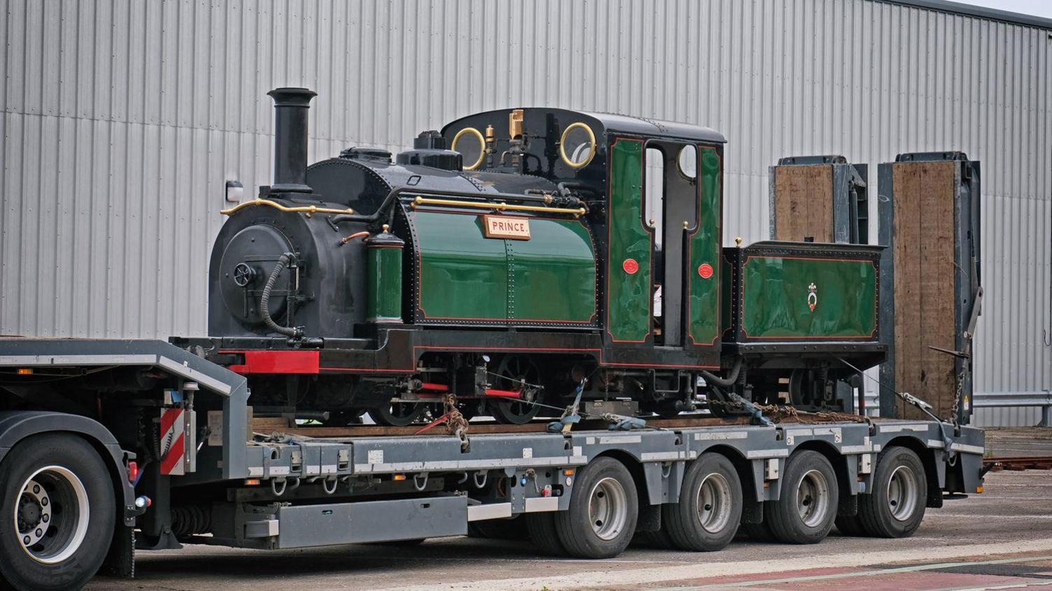 A vintage green steam locomotive named "Prince" is loaded on a flatbed trailer parked beside a large industrial building
