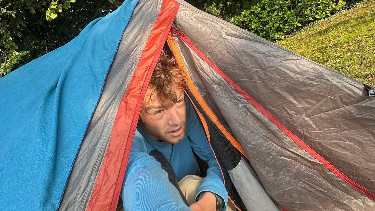 Ollie, looking tired as though he has just woken up, pokes his head out of a blue, grey and red tent.