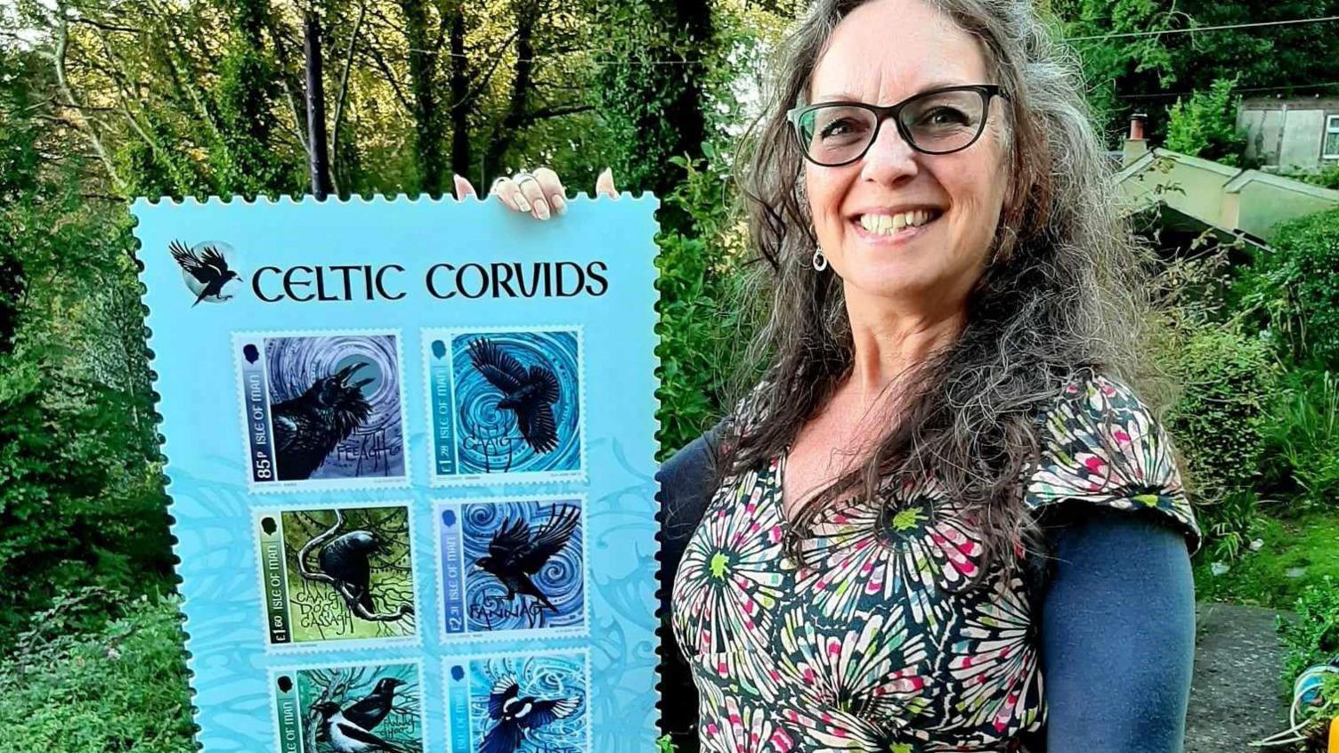 Julia Ashby Smith smiling while holding an large version of the stamps. She has curly hair and is wearing glasses.