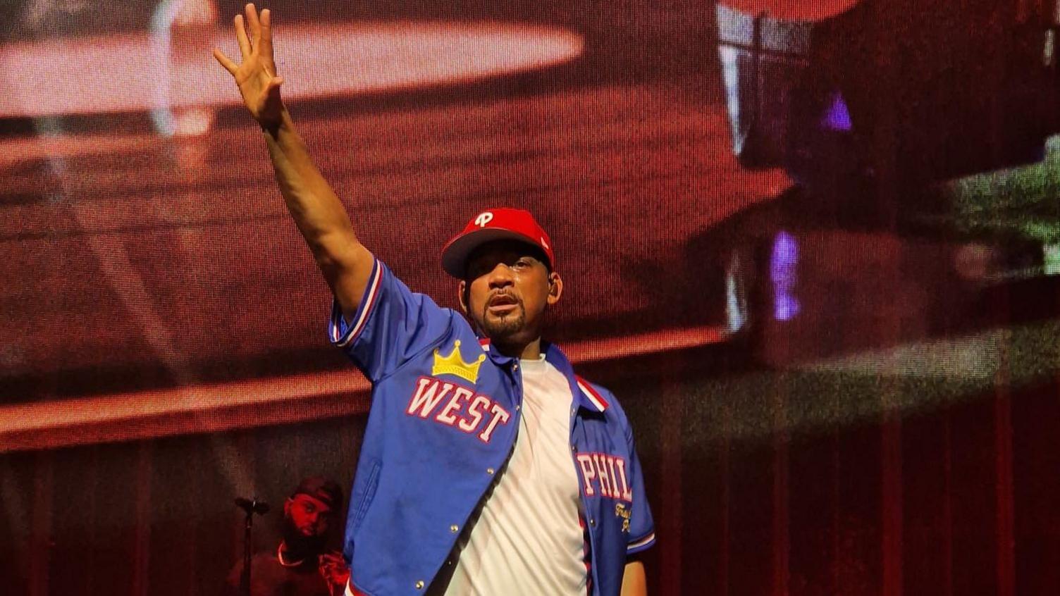 Will Smith in a white T-shirt and open blue baseball shirt with West Phil on it, and wearing a red baseball cap slightly twisted to the right. He is on stage and has his right hand raised in the air. He looks a little out of breath and sweaty. The stage behind him is backlit in red.