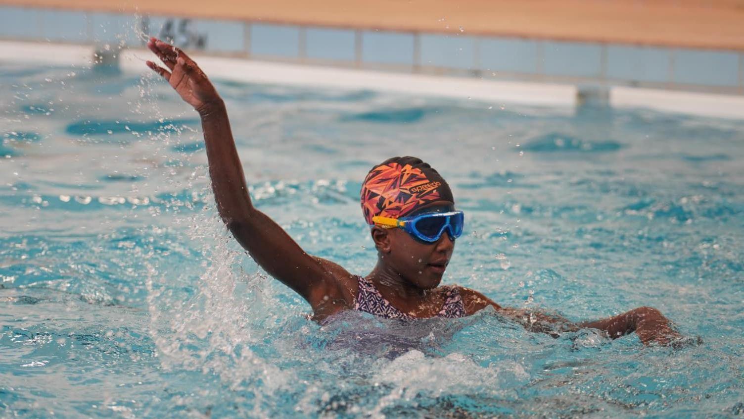 A girl wearing a pink and orange swimming hat and blue goggles is in a swimming pool. She is holding one arm above her out of the water. The edge of the swimming pool is in the background.