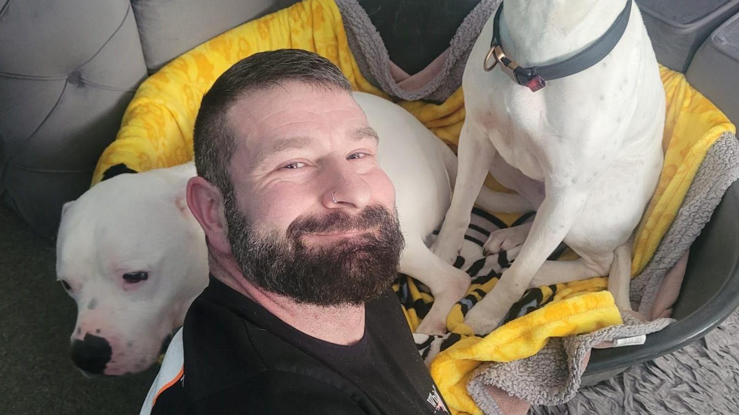 Anthony Webb smiles with his dog, Major seen on the left in a dog bed. The legs of the other side belong to his other dog Lexi. There is a yellow blanket in the dog bed. Anthony has short black hair with a black beard and a silver nose piercing in his right nostril.