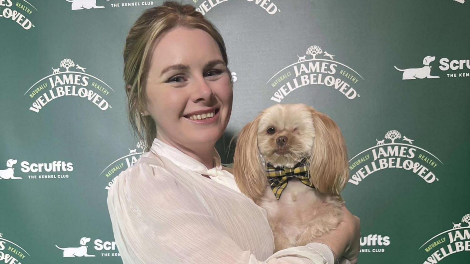 A woman smiling in a white shirt, holding a one-eye shih-tzu dog wearing a Cornish bow tie, in front of a board saying 'Scruffts'