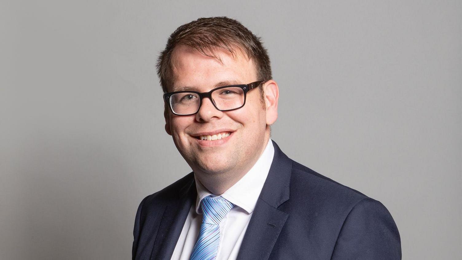 Young man with glasses in a navy suit jacket, white shirt, blue tie, smiles 