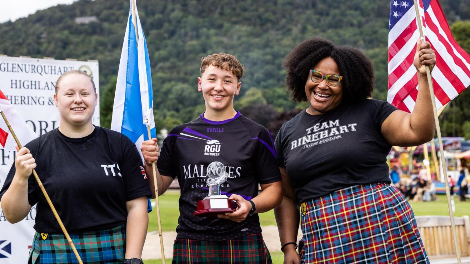Eve Robson, Juliet Ramsay and Gabriella Rinehart are standing side by side. Juliet is holding a Saltire flag and her winner's trophy. Eve and Gabriella are also holding the flags of their countries. The women are smiling. In the background is the Highland games arena and a tree covered hillside.