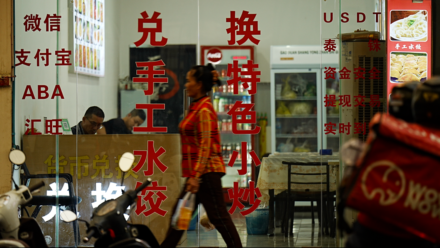 A woman walks down a road in front of the glass walls and doors of a business, with red writing in Cambodian, Chinese and Western scripts. Inside two men appear to work at a wooden desk while fridges with food and drink sit at the back of the store