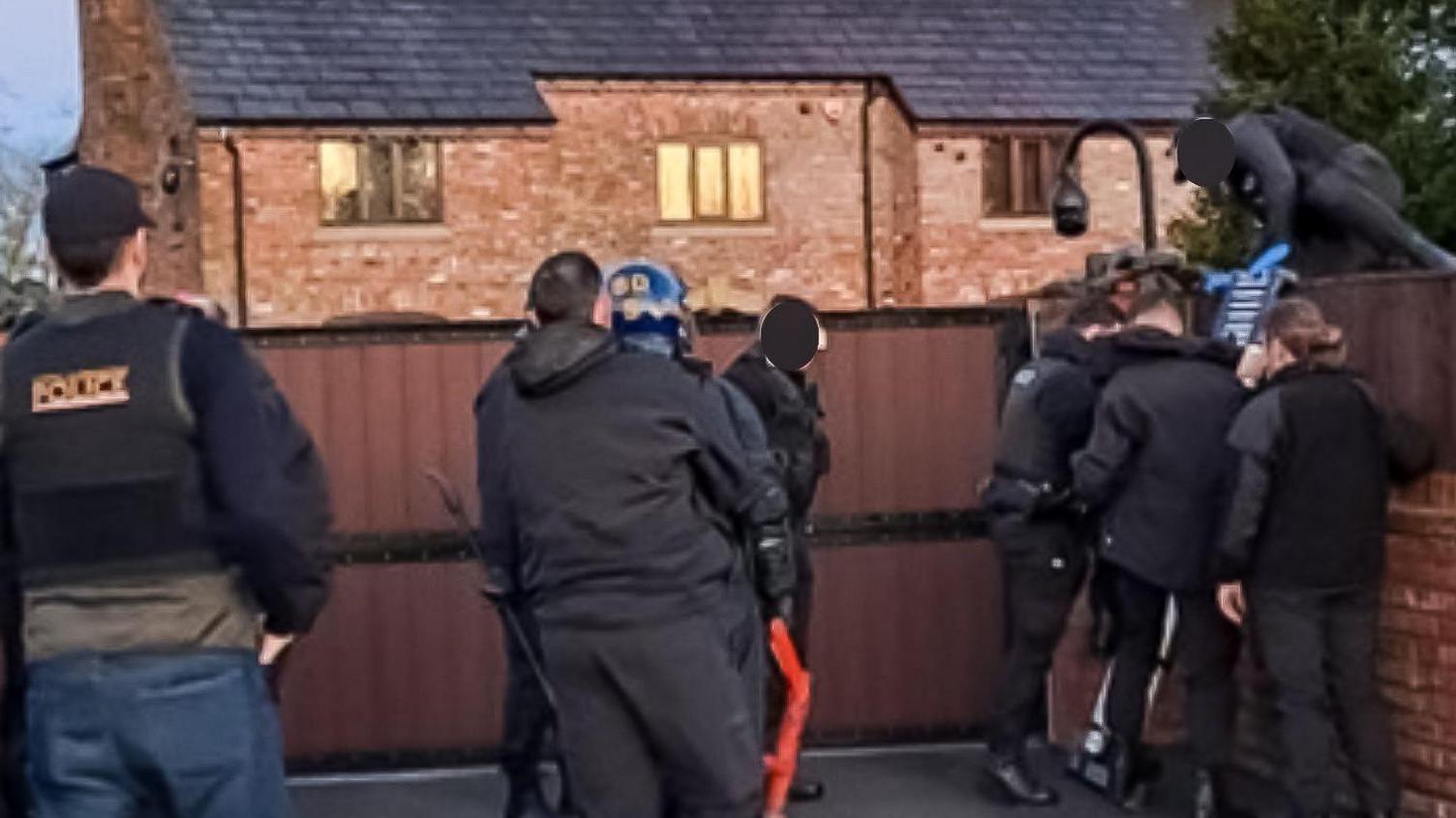 A group of police officers in black clothing, some wearing body armour, standing in the drive of a red brick house. Three officers and standing next to the wall one climbs over the fence from the top of a ladder. 