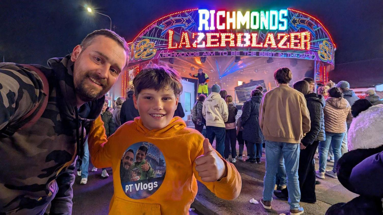 A man in a camouflaged jumper holding his arm out to take a selfie with his son, a young boy smiling and doing a thumbs up. He is wearing a yellow jumper that says PT Vlogs on it and has another selfie of him and his dad on it. Behind them are people stood around a neon sign which says Richmonds Lazerblazer.
