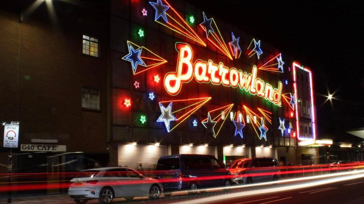 The Barrowland sign lit up. Cars are parked outside and a flare shows a car driving passed.