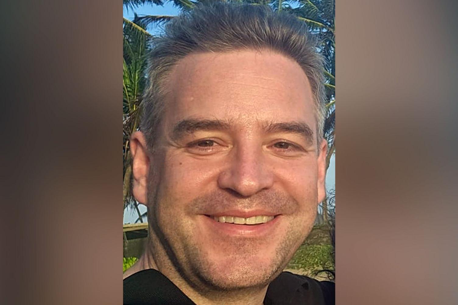 John Ames closeup of face, he has greyish hair, in the background are tropical trees.