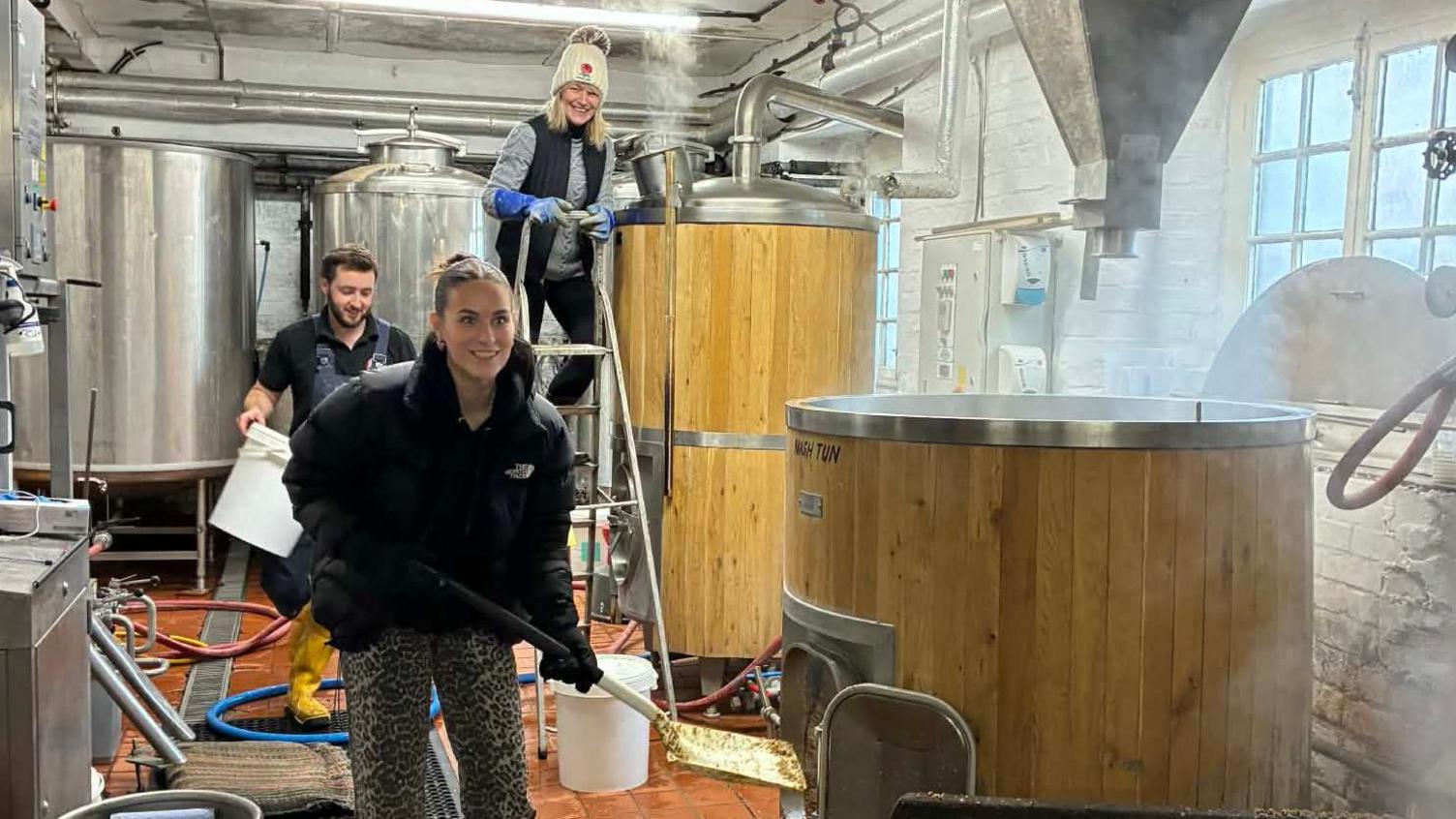 Georgina and Tina are in the brewery.  Tina is on top of a ladder next to a tank which has steam coming out of it. Georgina is holding a spade.