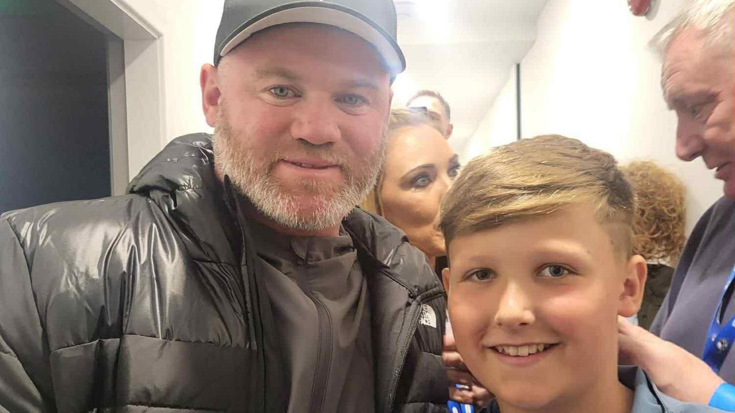 Mason is standing beside Wayne Rooney. He is wearing a tracksuit bottom top. Wayne is wearing a great tracksuit and black cap.