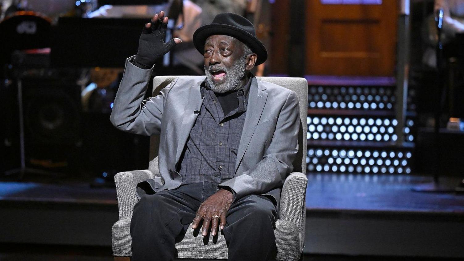 Garrett Morris sits on a light grey armchair holding a hand in a fingerless glove up. He wears a black hat, grey jacket and black trousers