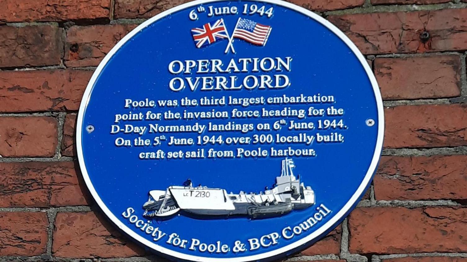 A round blue plaque on a brick wall. It reads: Operation Overlord, Poole was the third largest embarkation point for the invasion force heading for the D-Day Normandy landings on 6th June 1944. On the 5th June 1944 over 300 locally built craft set sail from Poole Harbour.