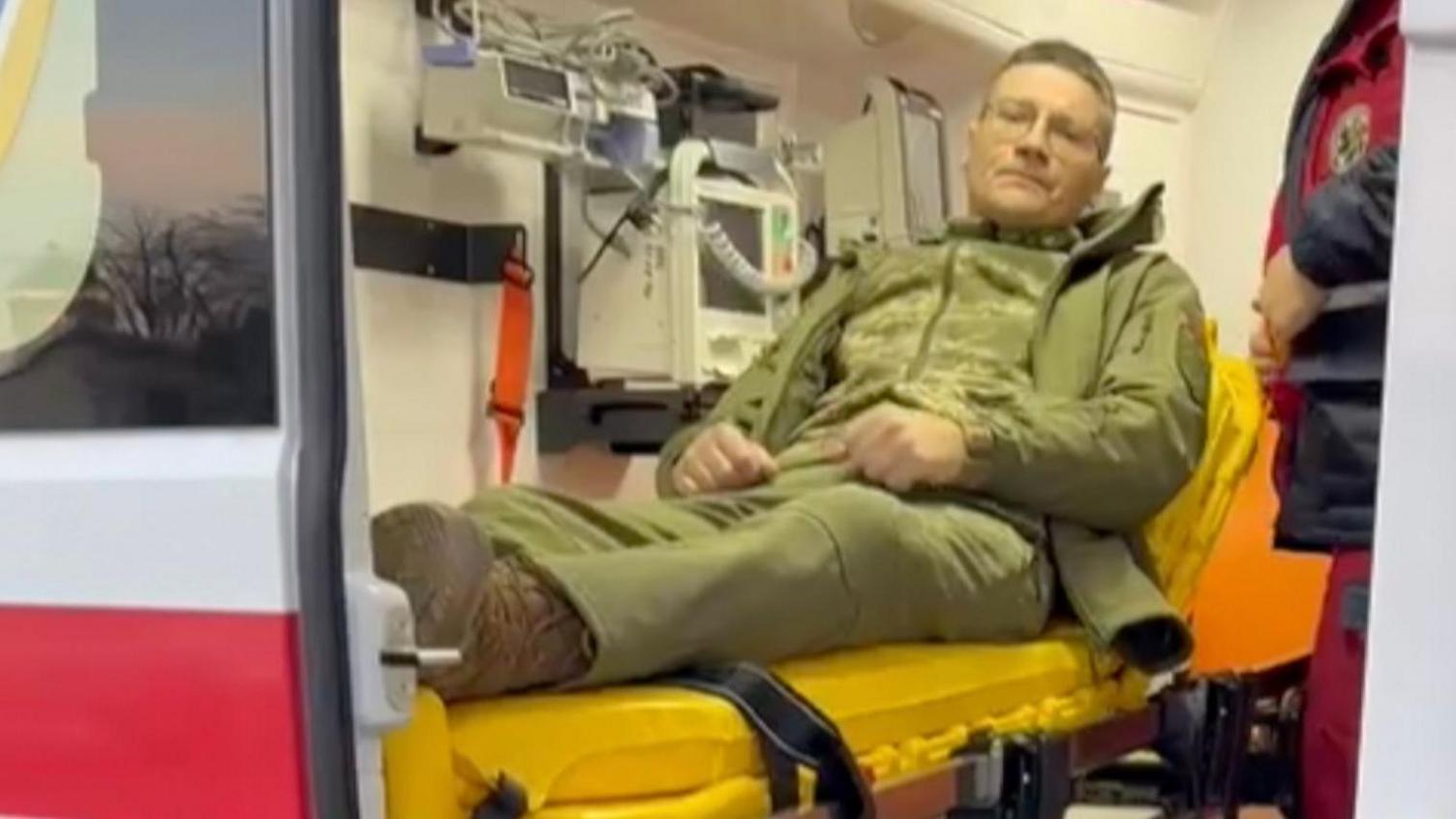 A man dressed in army khaki green uniform, lies on an yellow stretcher inside an ambulance. The man is sitting up and is wearing glasses.