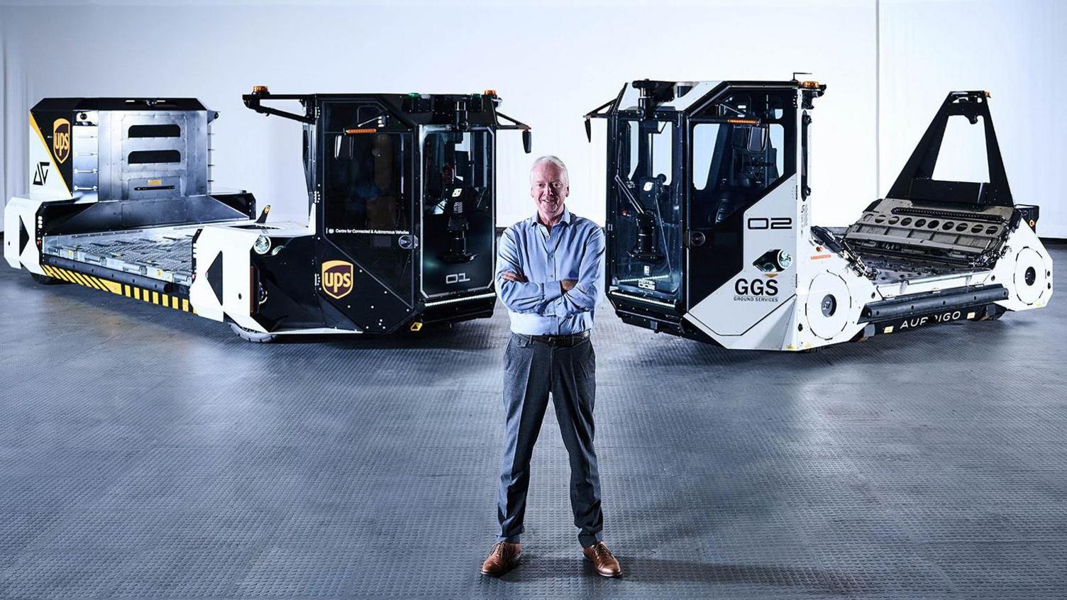 A man standing an a dark warehouse that has dim lighting. He is standing with arms crossed in front of two self-driving vehicles that are used to move airport baggage. He is wearing suit trousers and a blue shirt and has short grey hair. He is smiling.
