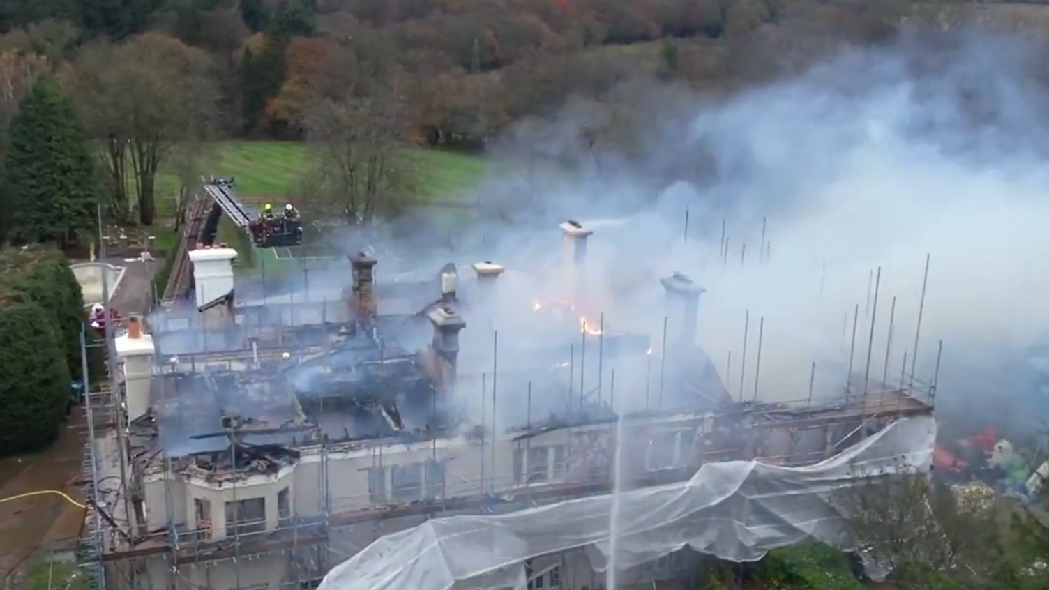 An aerial view of Headley Hall, showing smoke rising from the fire, with flames still burning. Water jets from fire engines are being trained on the roof.