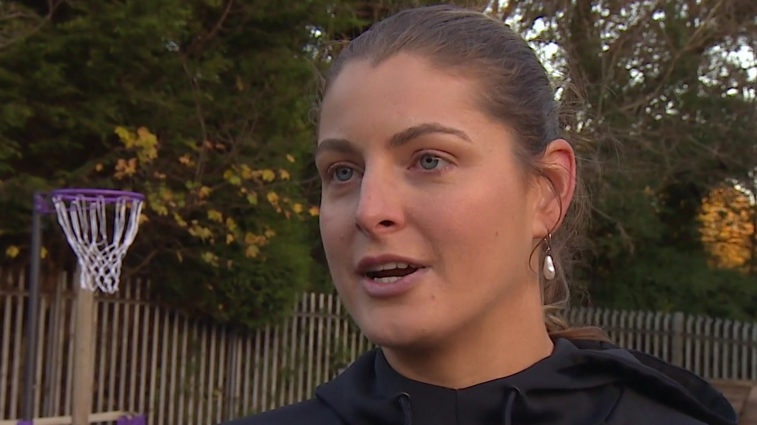 Gabby is standing outside on a court with a netball hoop on the left of the image. Her brown hair is tied back and she has pearl earrings in.