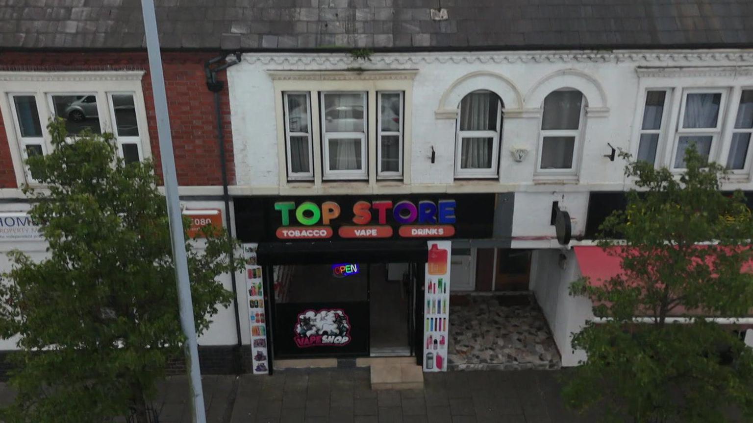 An aerial view of Top Store on Nantwich Road in Crewe. The shop is below a row of residential flats, with trees on the left and right of the shot. There is a multi-coloured sign advertising the shop with Tobacco Vape and Drinks beneath it.