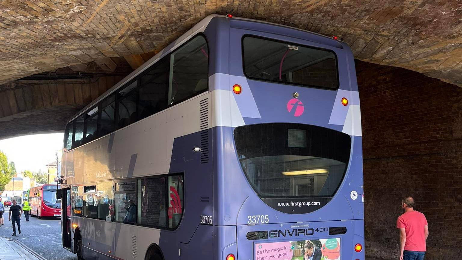 A purple and white blue double-decker bus stuck under the arch of a large bridge.