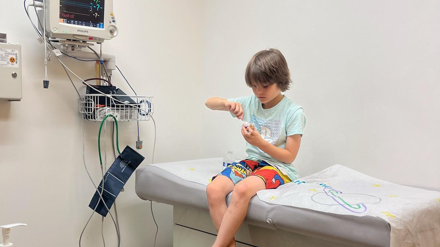 A boy in a medical room wearing shorts and a t-shirt while eating pudding on a bed.  
