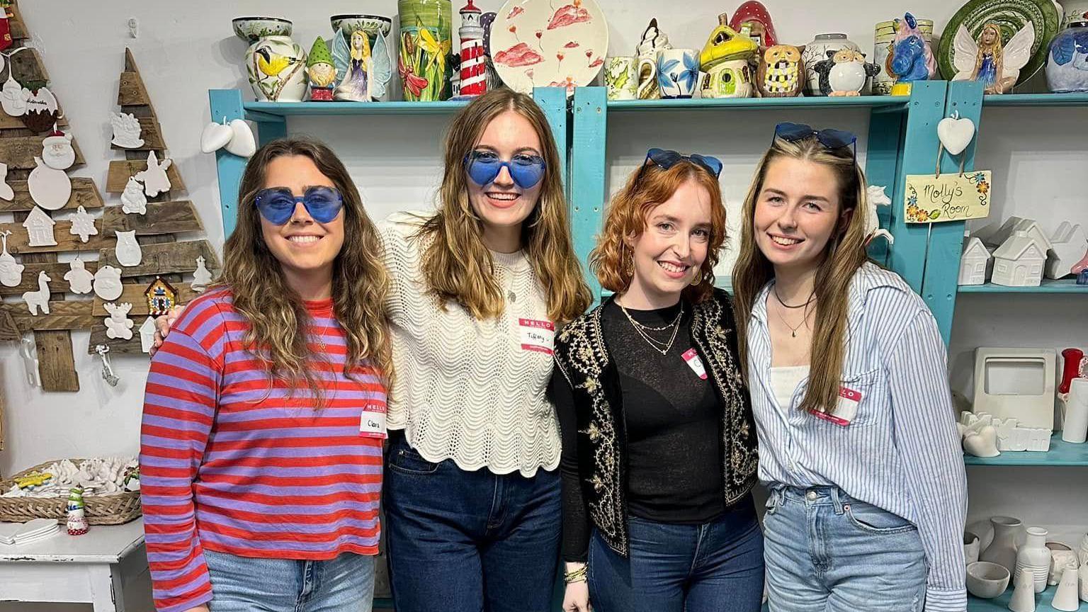 Social Girls Club volunteers stand in front of pottery on shelves. They are wearing brightly coloured clothes and blue love heart-shaped glasses and smiling. 