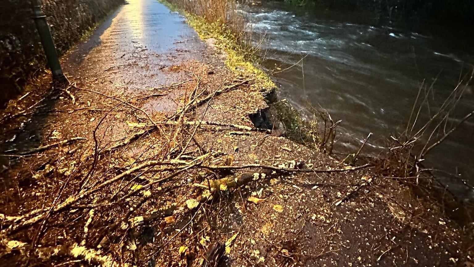 Branches on the pathway on the left with the river to the right. There is a chunk of pathway missing from the damage. 