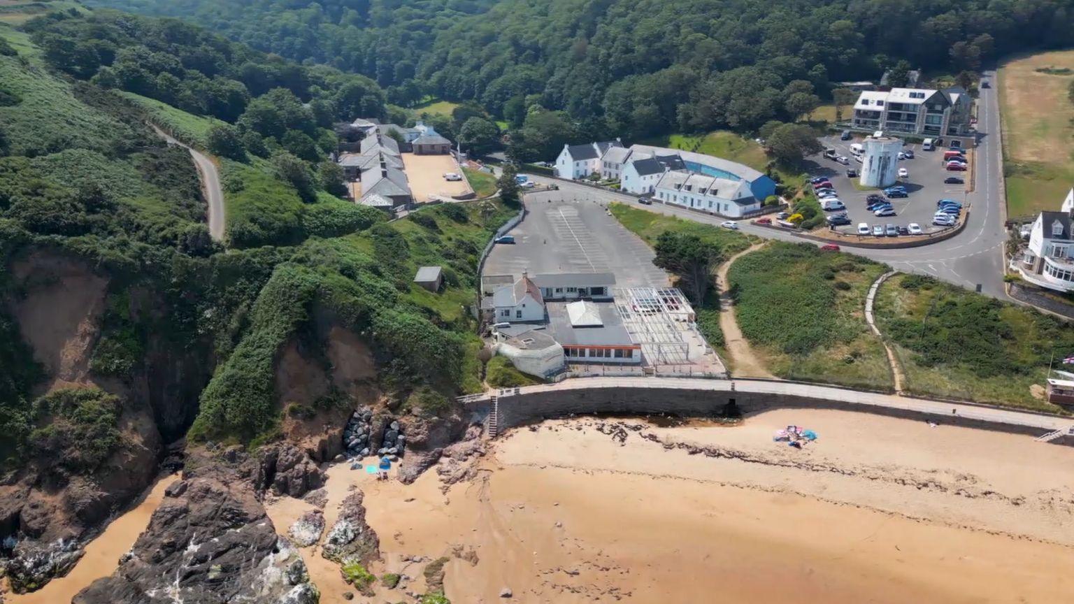 A drone photo of Greve De Lecq beach. At the bottom of the photo is the beach with browny-white sand, and on the left the sand turns into brown cliffs with green grass and hedges on top of them. There is also a car park and conrete.