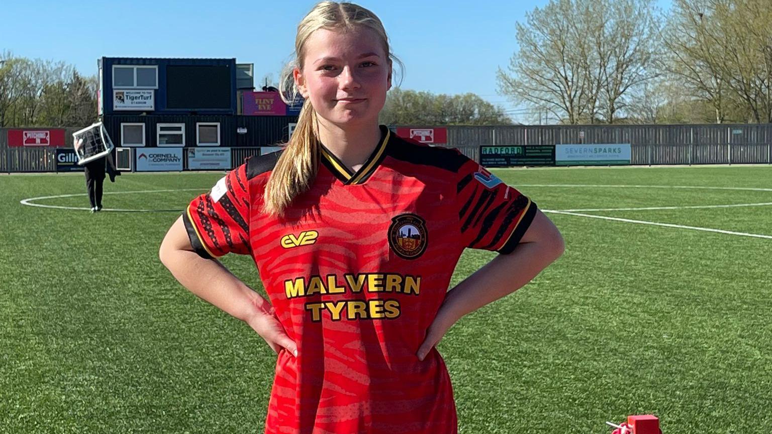 Imogen wearing a red football top standing in a football pitch with her arms on her waist and hair tied back, smiling at the camera.