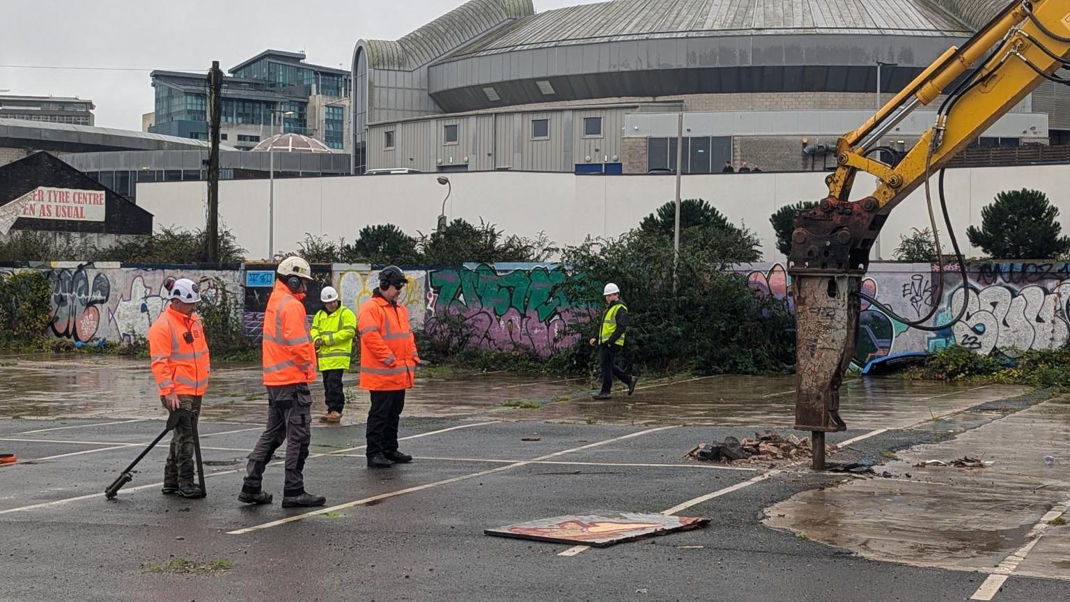 Workers in his-vis at the site. The workers are wearing his-vis jackets, helmets and ear defender. A heavy machine is digging into the concrete car park.