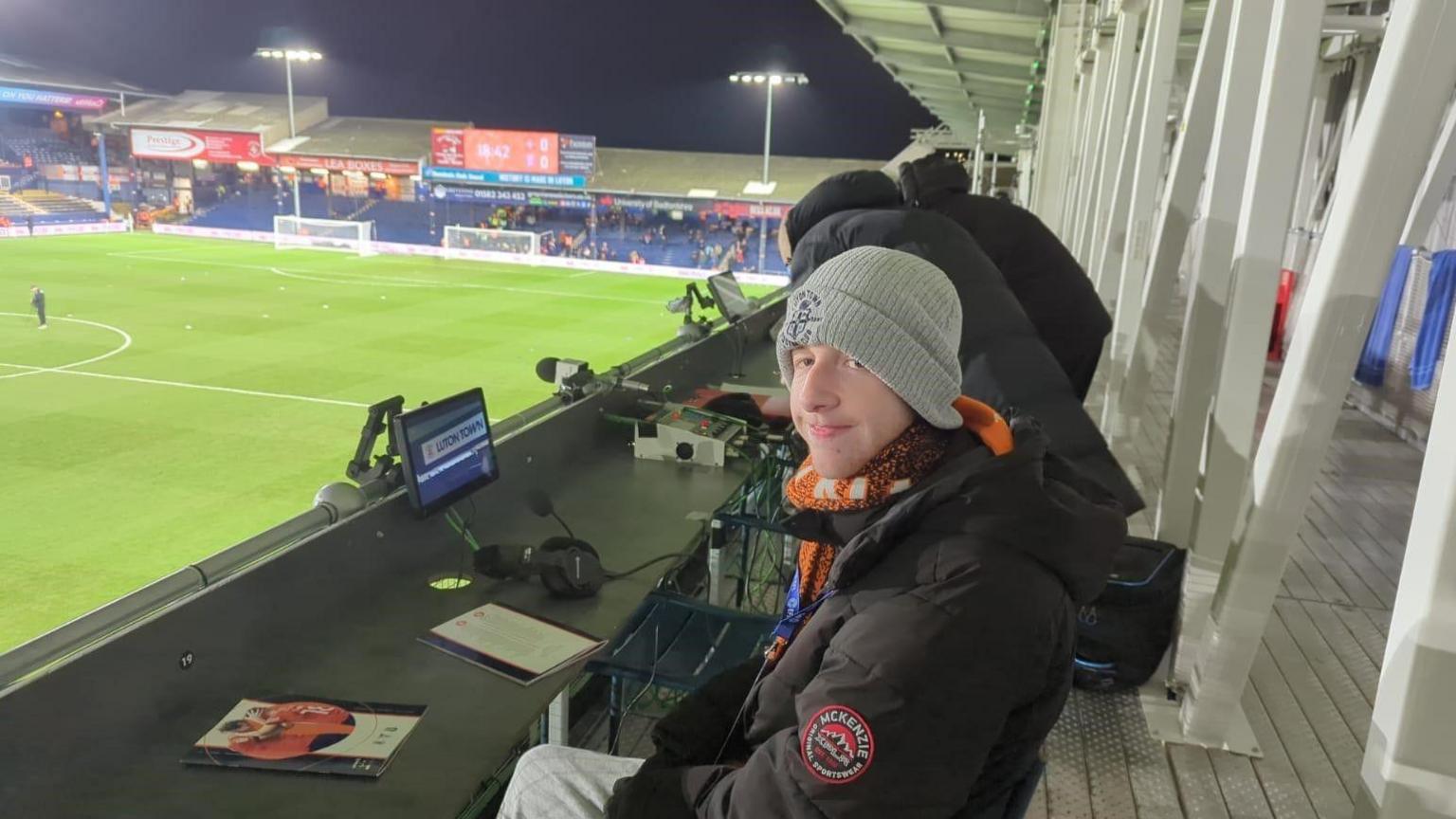 Aidan smiling at the camera from the BBC commentary box at a football game. He is wearing a grey beany hat and the football pitch is visible in the background