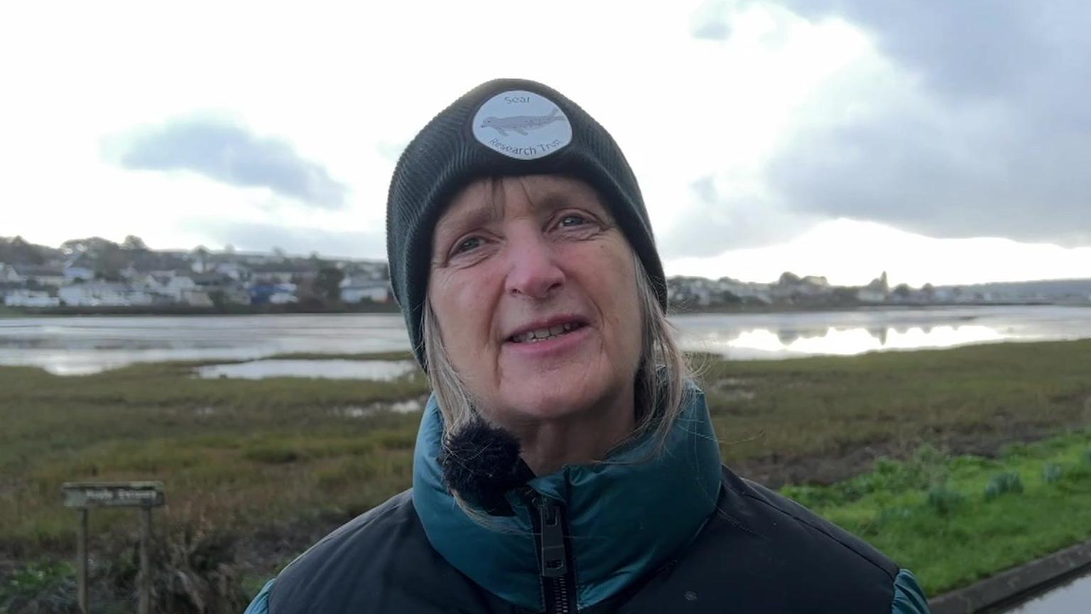 Sue Sayer stands in front of an estuary and marsh land area in Cornwall on a cloudy day while wearing a black beanie hat with a Seal Research Trust logo on it and a green and black coat.