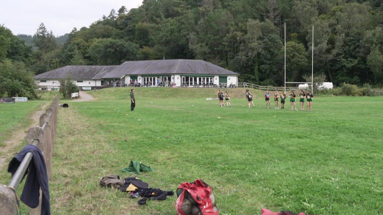 Chwaraewyr yn ymarfer ar gae Clwb Rygbi Nant Conwy. Mae adeilad y clwb yn y cefndir a rhan o'r coetir sydd wrth y safle.