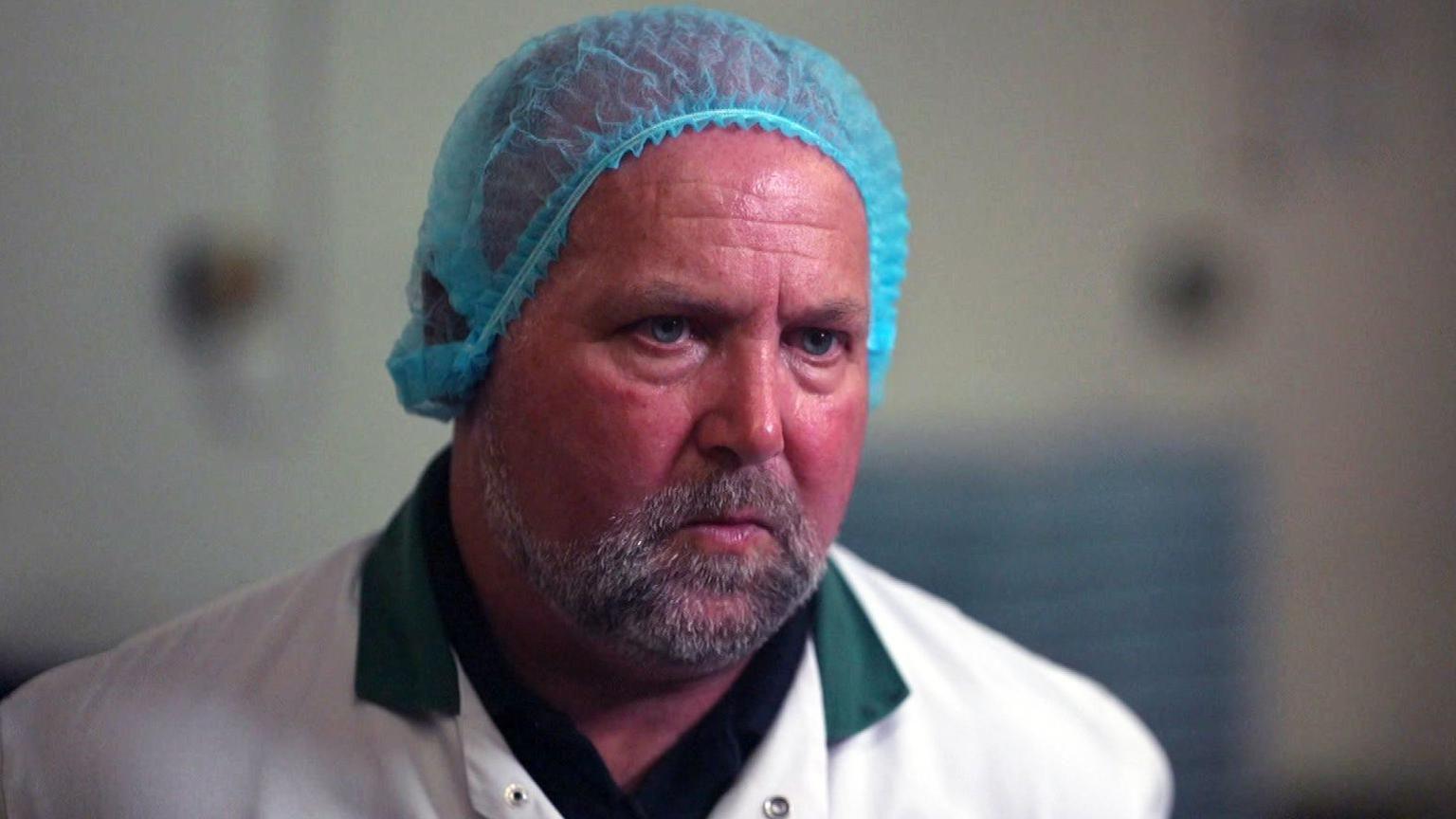 Allan Jones wearing a white lab coat over a black shirt and a blue hairnet. The background is out of focus but appears to be an indoor industrial or food production setting.

