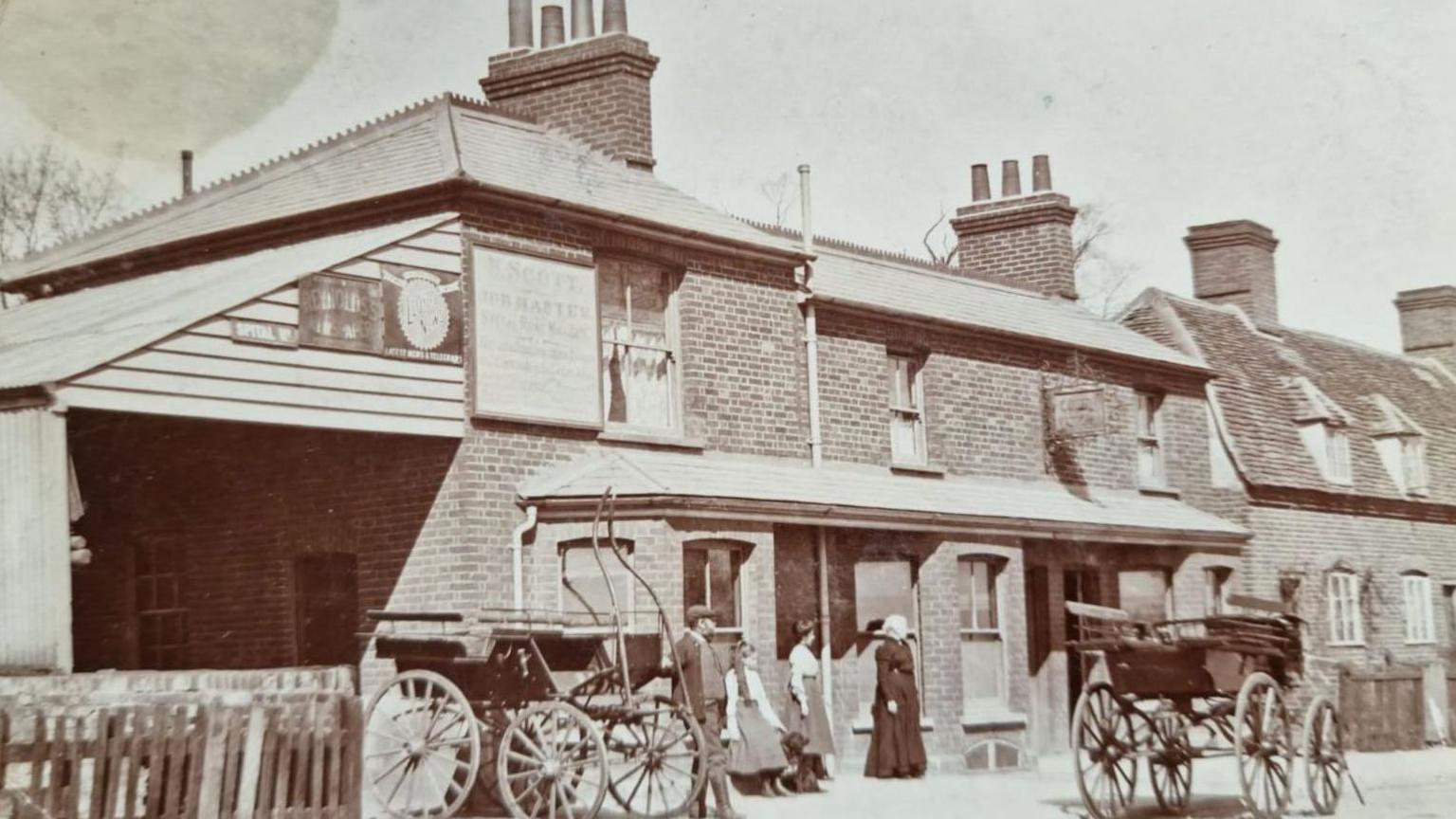 An old sepia coloured photo of the pub from outside. There are two horse drawn carriages but no horses. Four people can be seen stood outside in Victorian dress looking at a carriage.