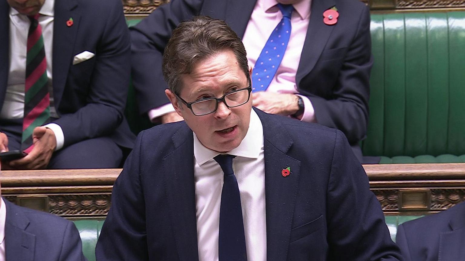 Alex Burghart speaking in the House of Commons. He has brown hair, wearing glasses, a navy blazer, white shirt and navy tie with poppy pin on his lapel. Other MPs are sitting on green leather benches beside and behind him.