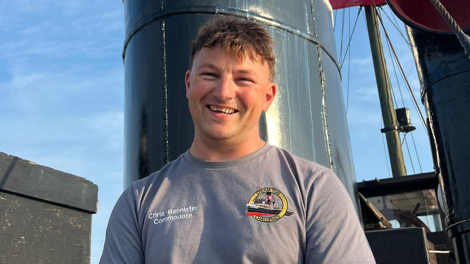 A smiling man, wearing a grey t-shirt with a logo of a vessel, is standing on a boat.