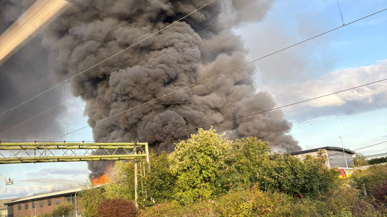 Plumes of dark grey smoke are billowing into the blue sky and some brewery buildings can be seen close by